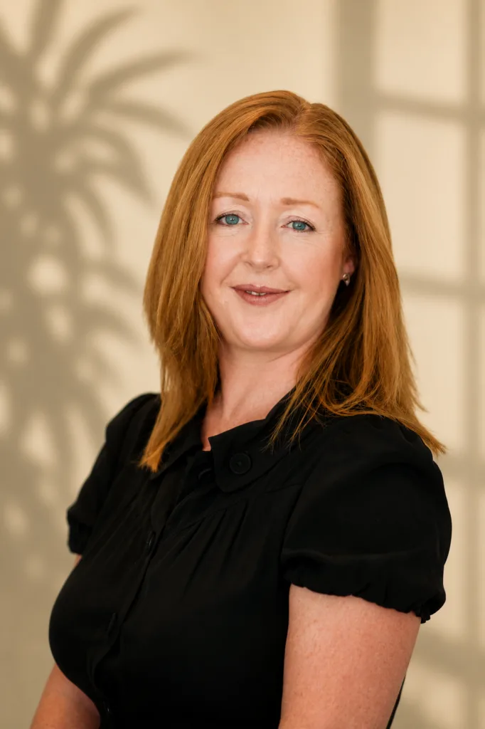 Red-haired woman wearing a black blouse poses against a softly lit neutral background