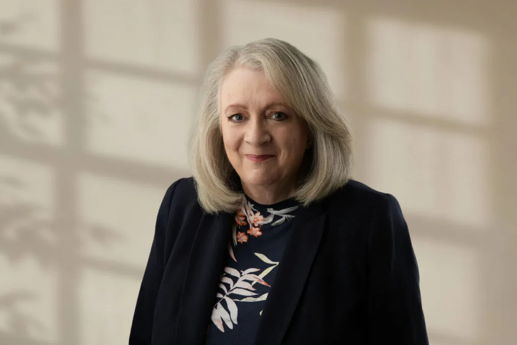 Woman with shoulder-length grey hair wearing a dark blazer and floral top against a blurred neutral background