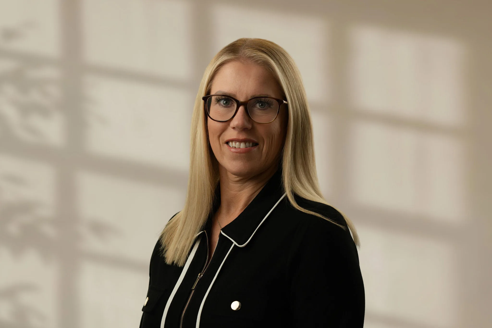 Blonde woman wearing glasses and a black jacket with white piping smiling at the camera