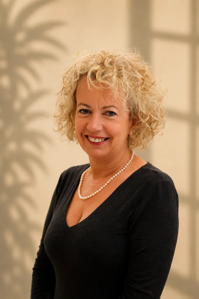 Woman with curly blonde hair wearing black top and pearl necklace against beige background shadow