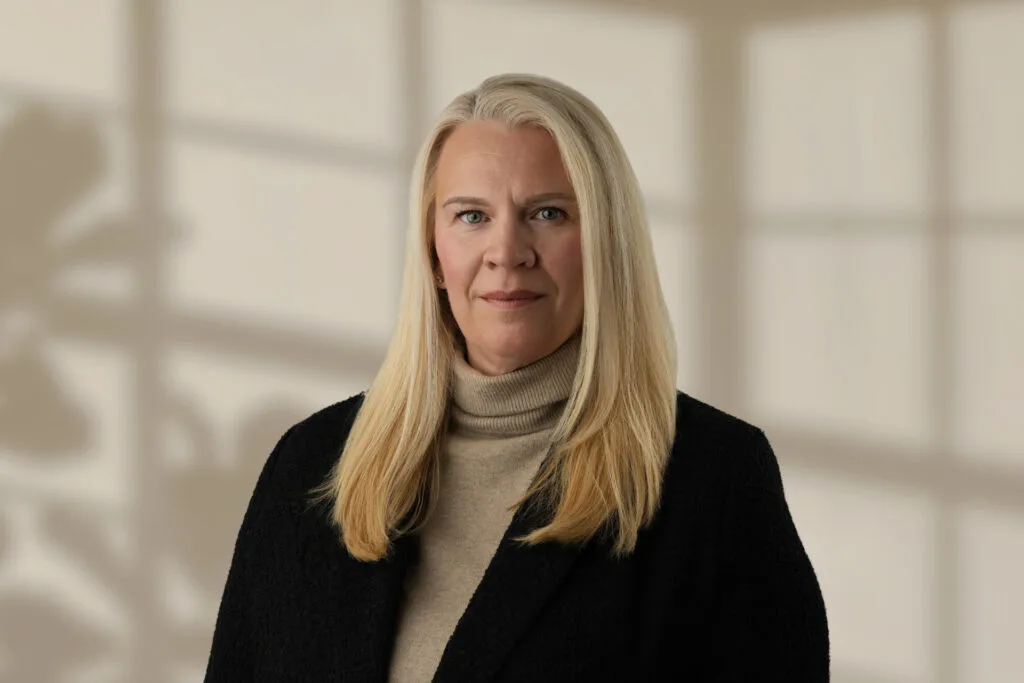 Woman with long blonde hair wearing a beige turtleneck and black jacket against blurred background