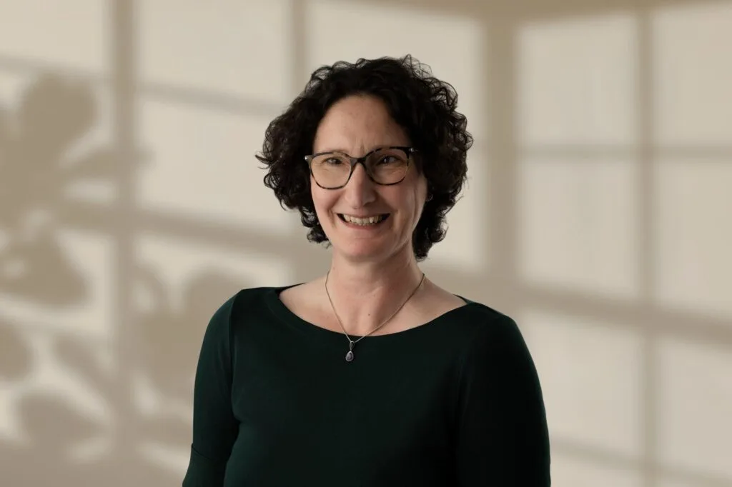 Mary Lomas with short curly dark hair, glasses, and green top against beige background