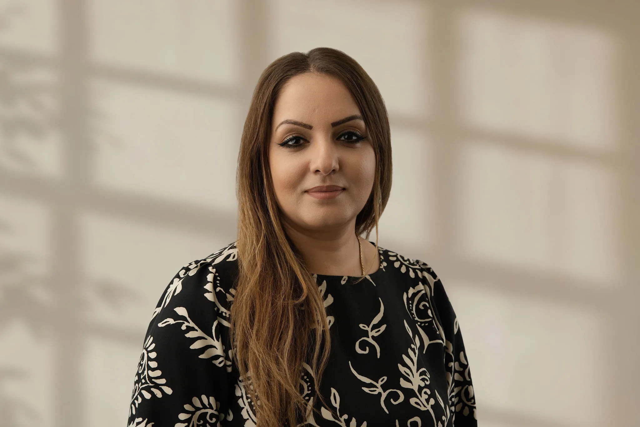 Woman with long brown hair wearing patterned black top smiling indoors with soft light