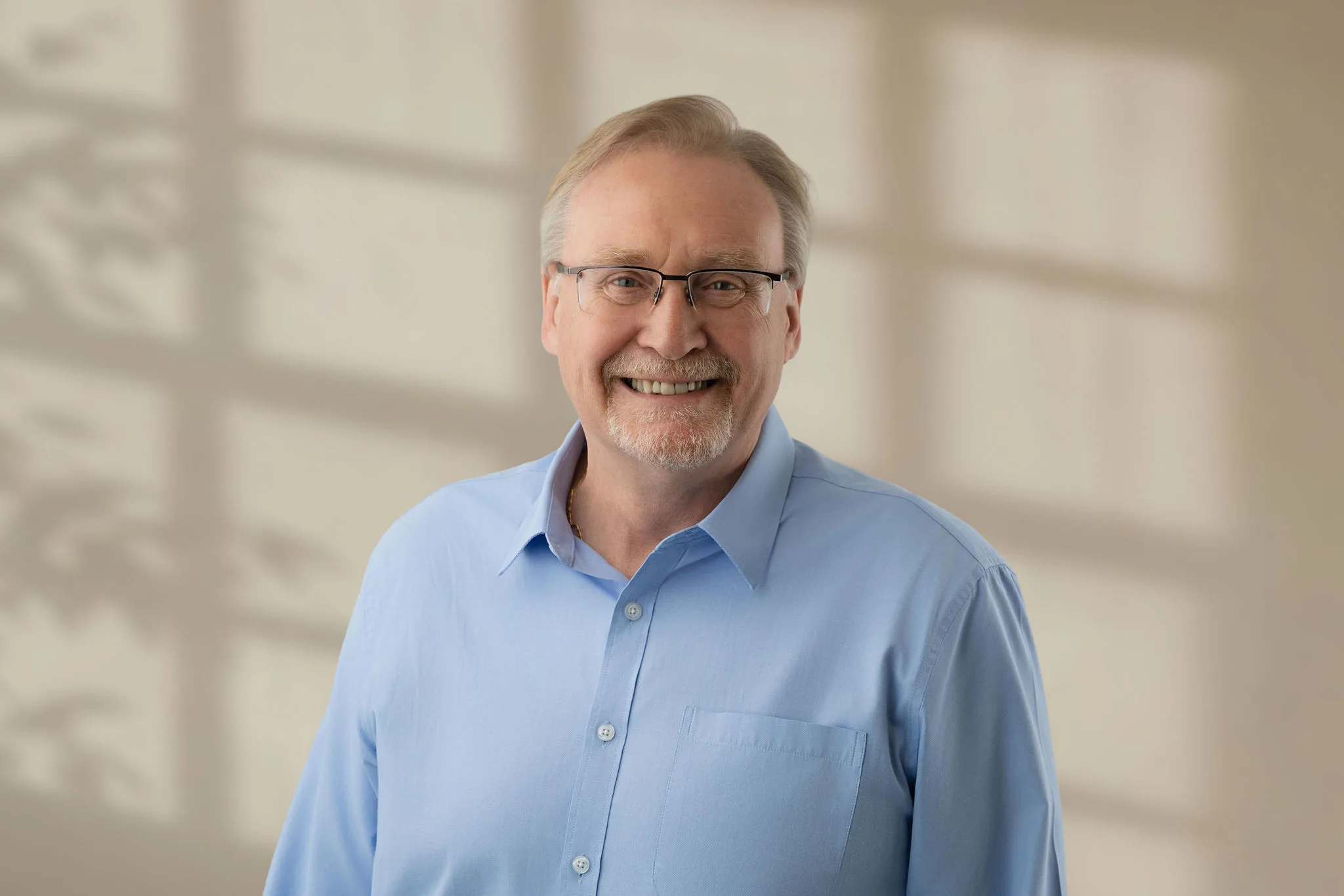 Older man with glasses and light beard smiling in blue shirt with soft room lighting
