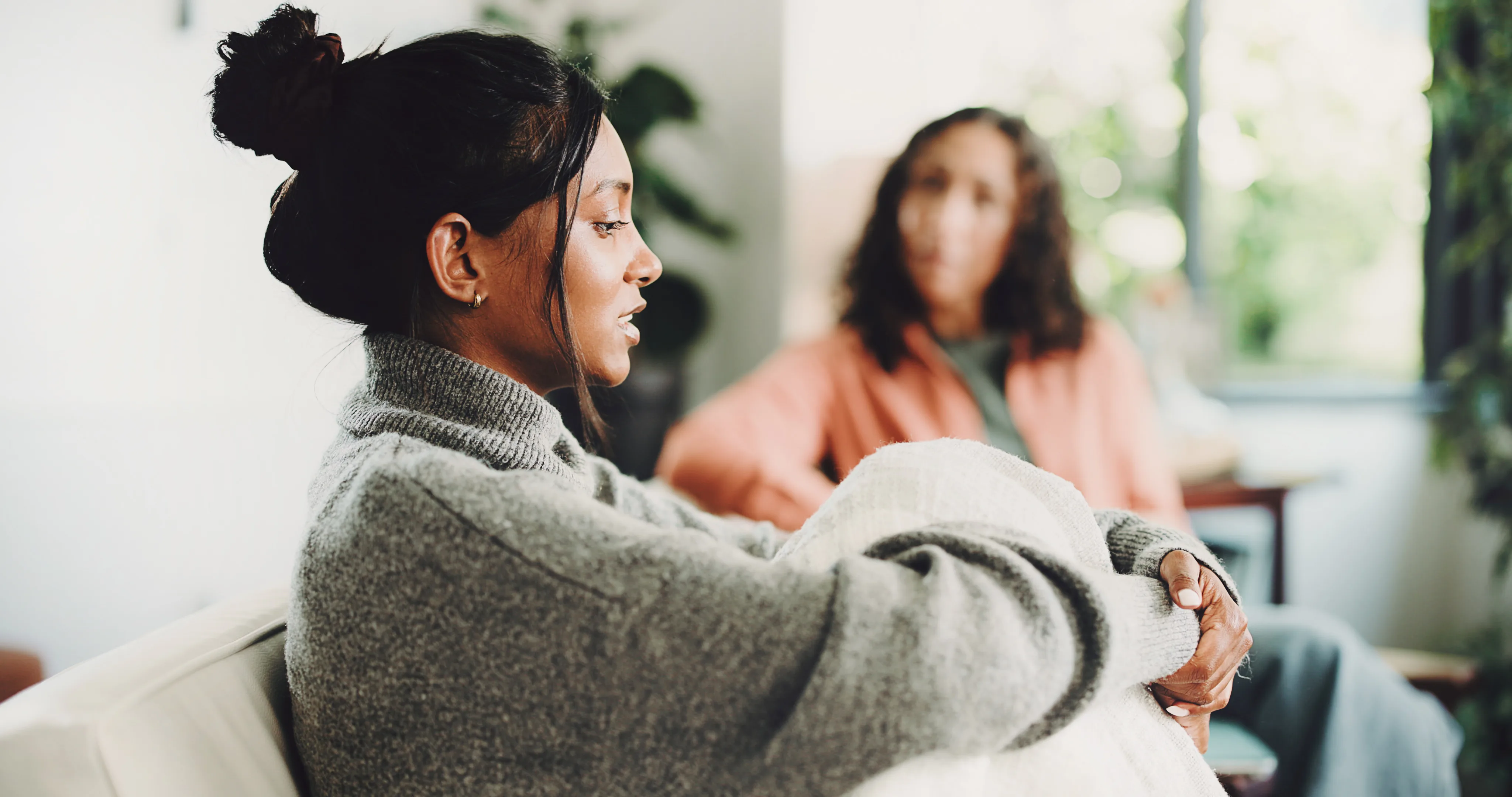 Woman in grey sweater sitting on sofa talking to another woman out of focus