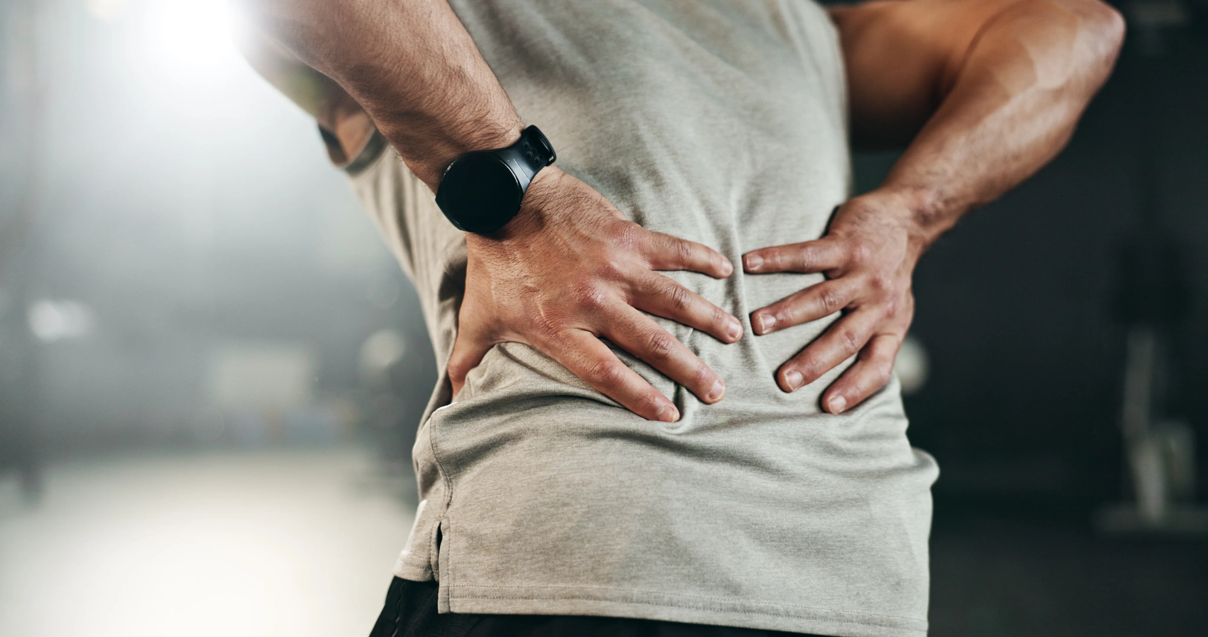 Man holding his lower back, seen from behind, wearing a watch and grey t-shirt