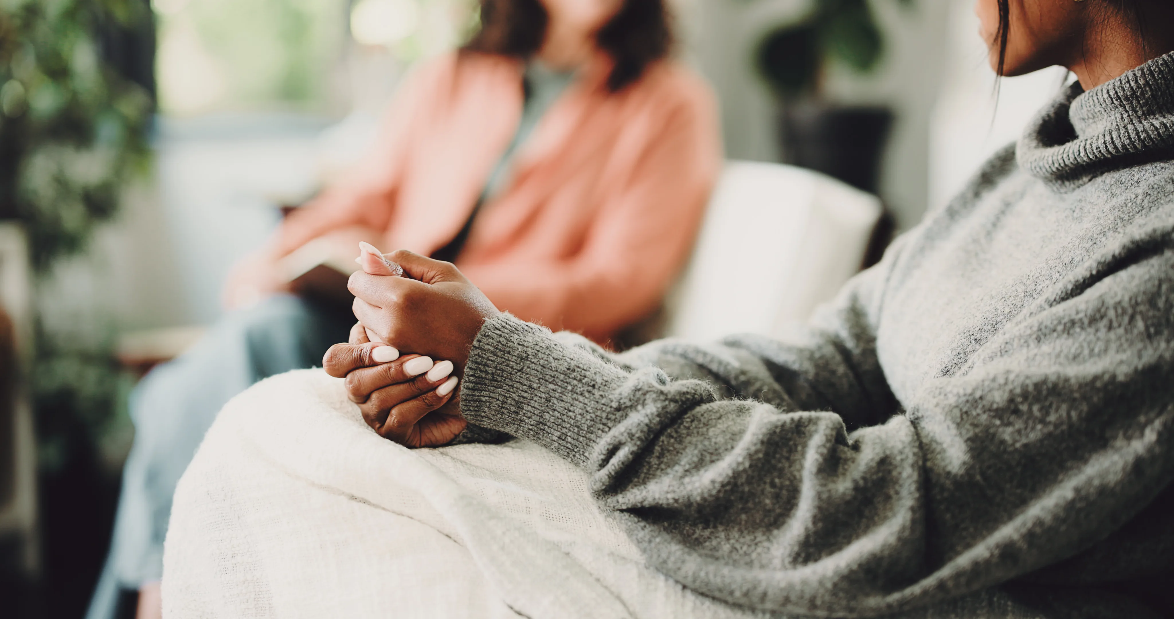 Woman wearing grey sweater sitting with hands clasped on lap, blurred person in background