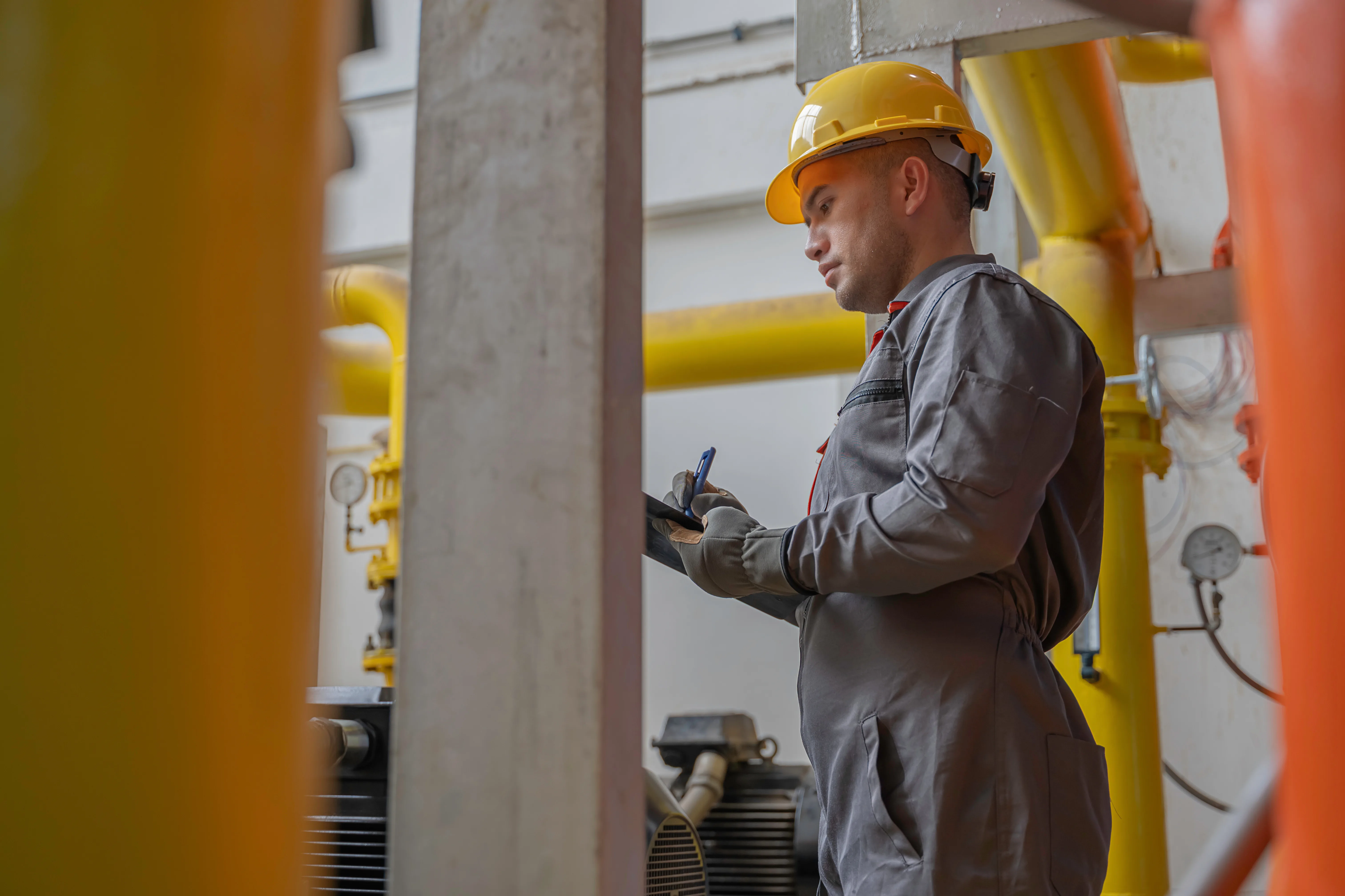 Man in yellow hard hat and grey coveralls writing on clipboard near yellow piping