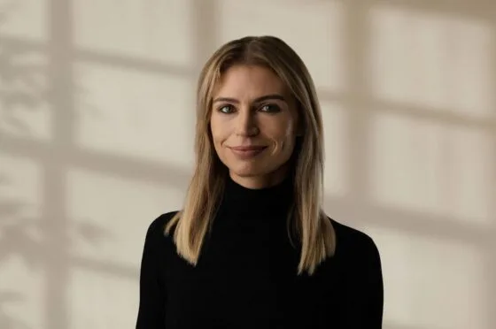 Alex Lynch wearing a black turtleneck sweater against a beige background
