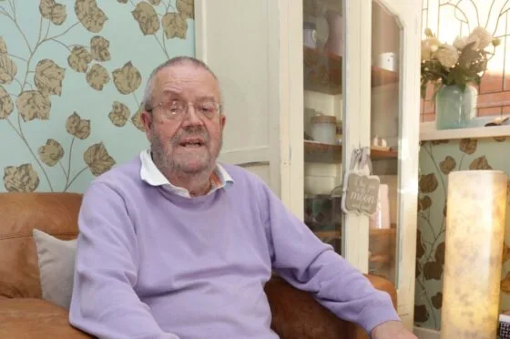 Older man wearing glasses and purple sweater sitting on brown leather sofa indoors