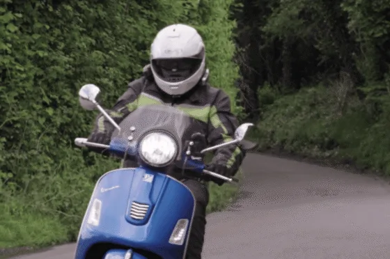 Person wearing helmet and protective clothing riding blue motor scooter on narrow road