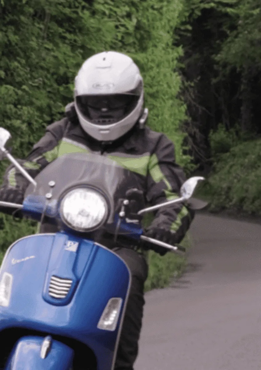 Person wearing helmet and protective clothing riding blue motor scooter on narrow road