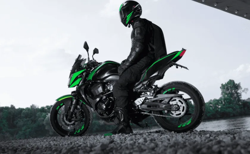Motorcyclist in black gear and helmet sitting on a black and green motorcycle near a river under a structure