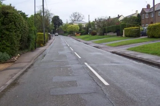 Empty residential street lined with houses and grassy gardens under cloudy sky