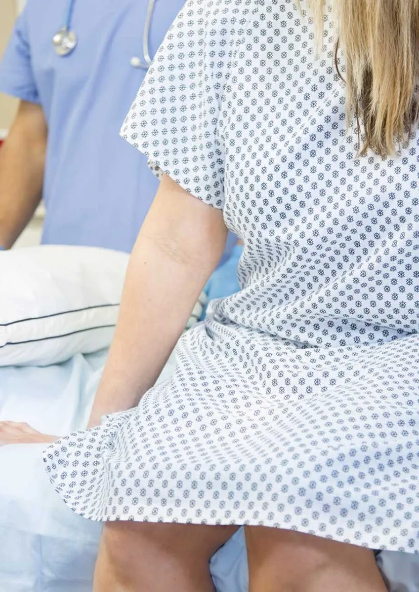Woman in patterned hospital gown sitting on bed with healthcare professional in blue scrubs