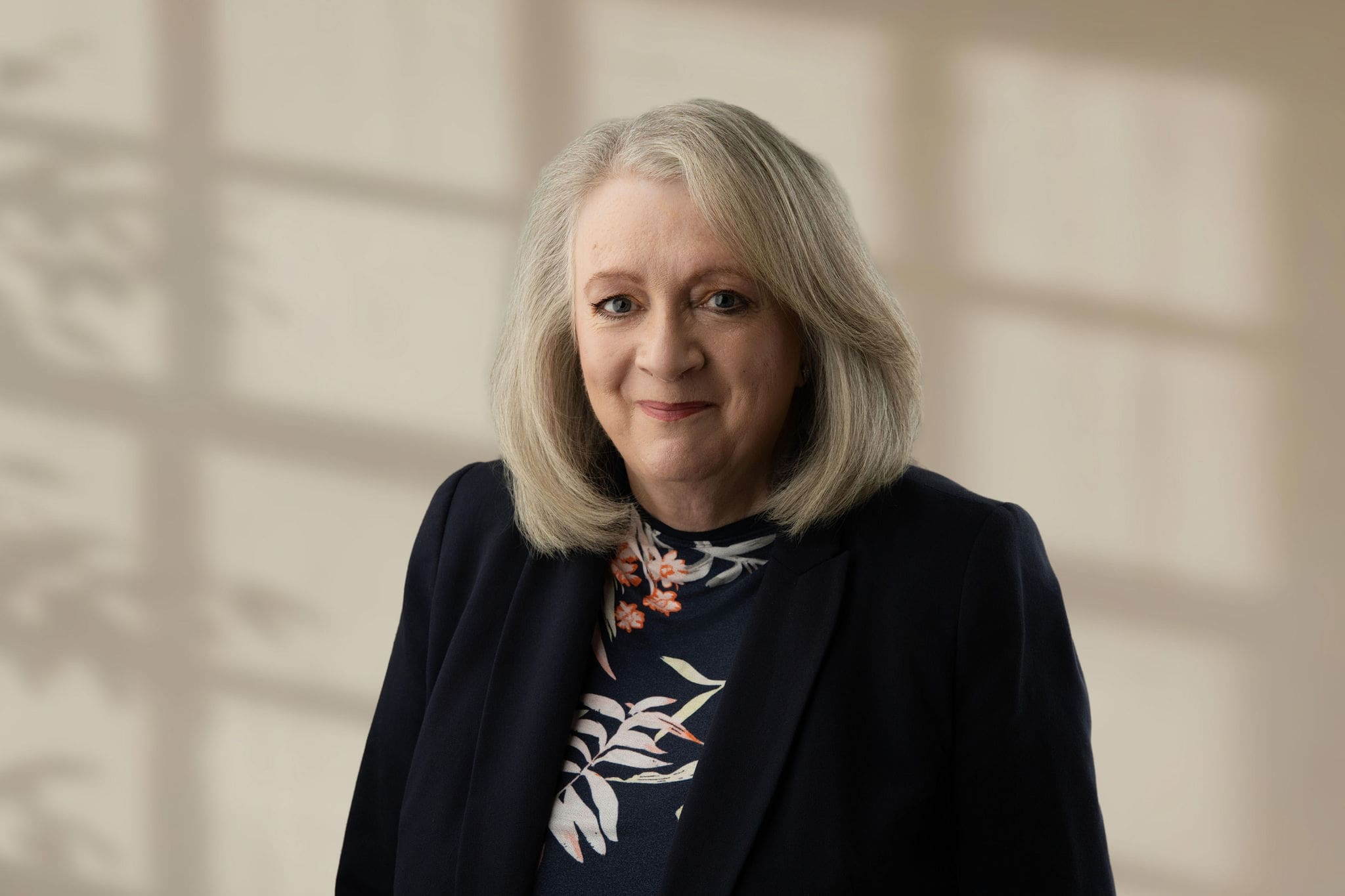 Woman with shoulder-length grey hair wearing a dark blazer and floral top against a blurred neutral background