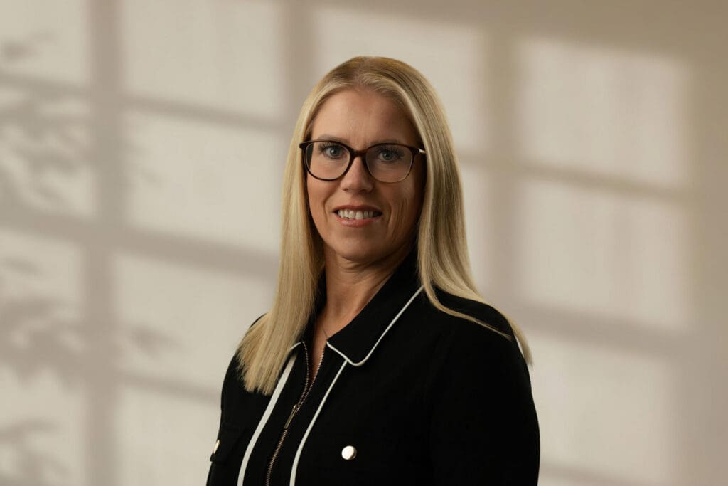Blonde woman wearing glasses and a black jacket with white piping smiling at the camera