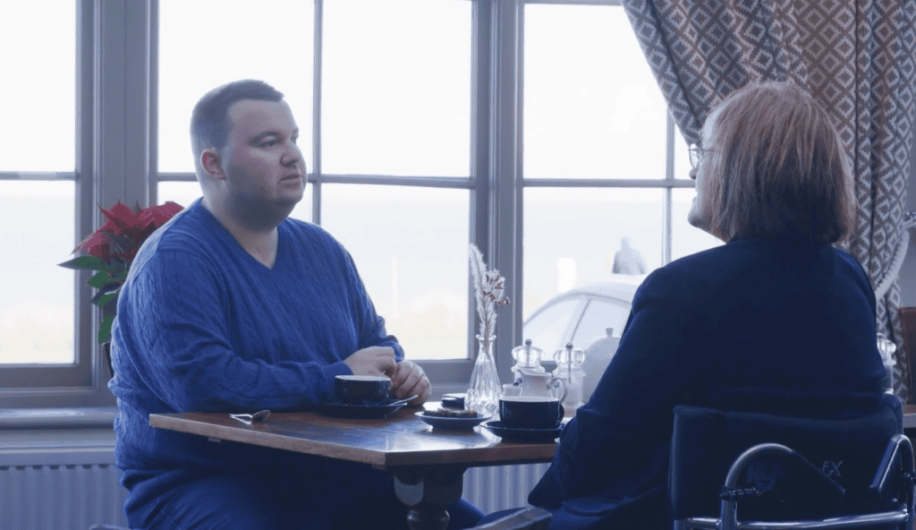 Two people sitting at a table by a window, with coffee cups and a vase between them
