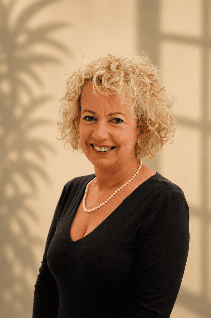Woman with curly blonde hair wearing black top and pearl necklace against beige background shadow