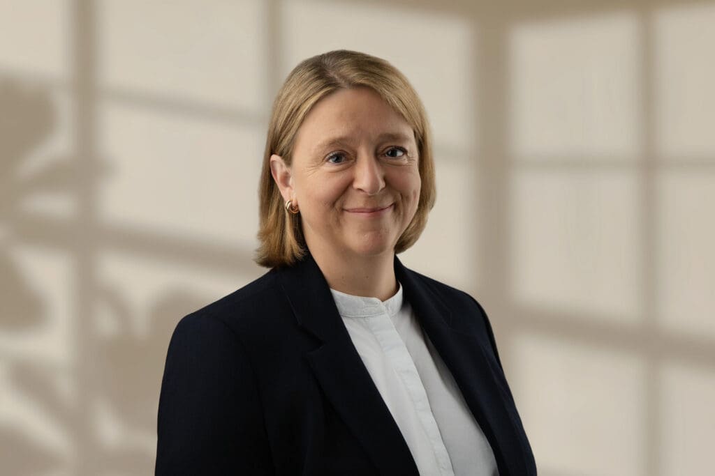 Jill Heywood wearing a dark blazer and white blouse against a beige background
