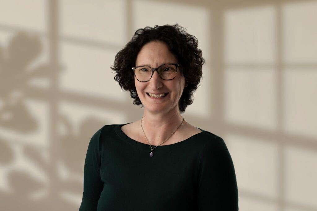 Mary Lomas with short curly dark hair, glasses, and green top against beige background