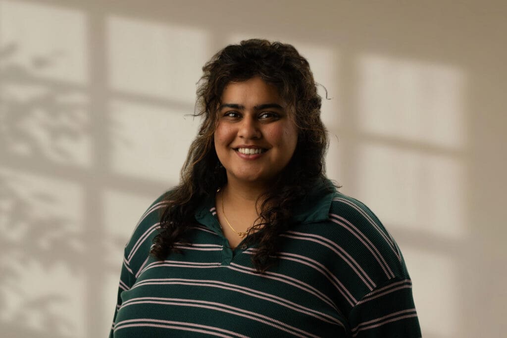 Nermeen Salahuddin smiling in a striped dark green and pink shirt before a blurred window shadow background