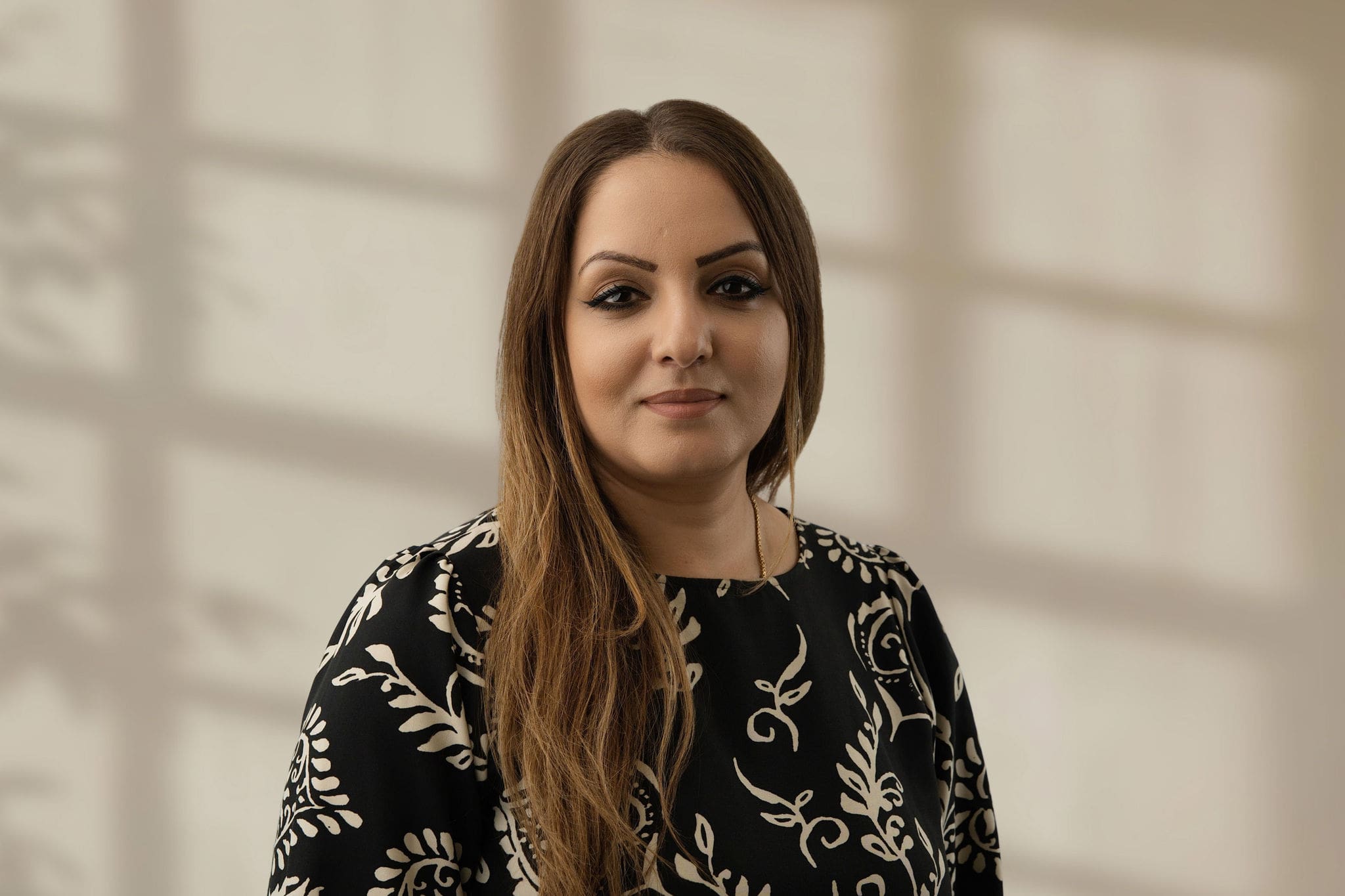 Woman with long brown hair wearing patterned black top smiling indoors with soft light