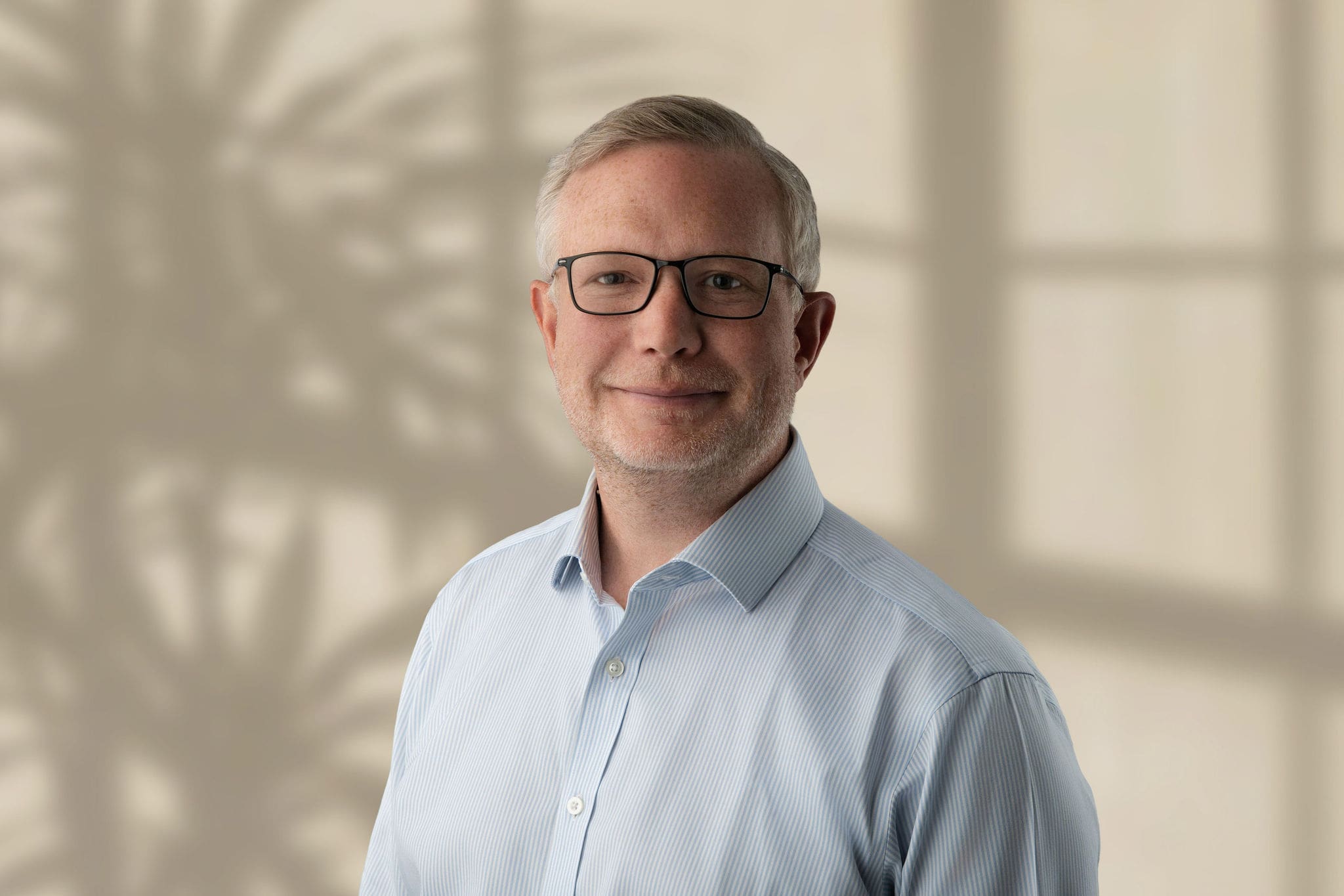 Man with light hair, glasses, and pale blue striped shirt against beige background shadow