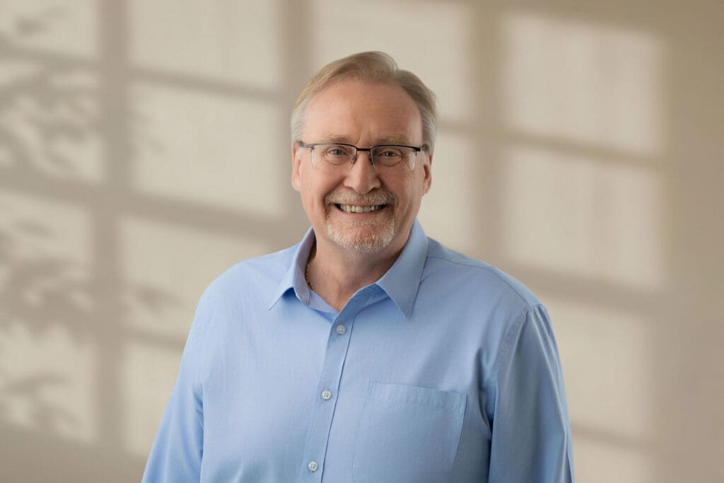 Older man with glasses and light beard smiling in blue shirt with soft room lighting