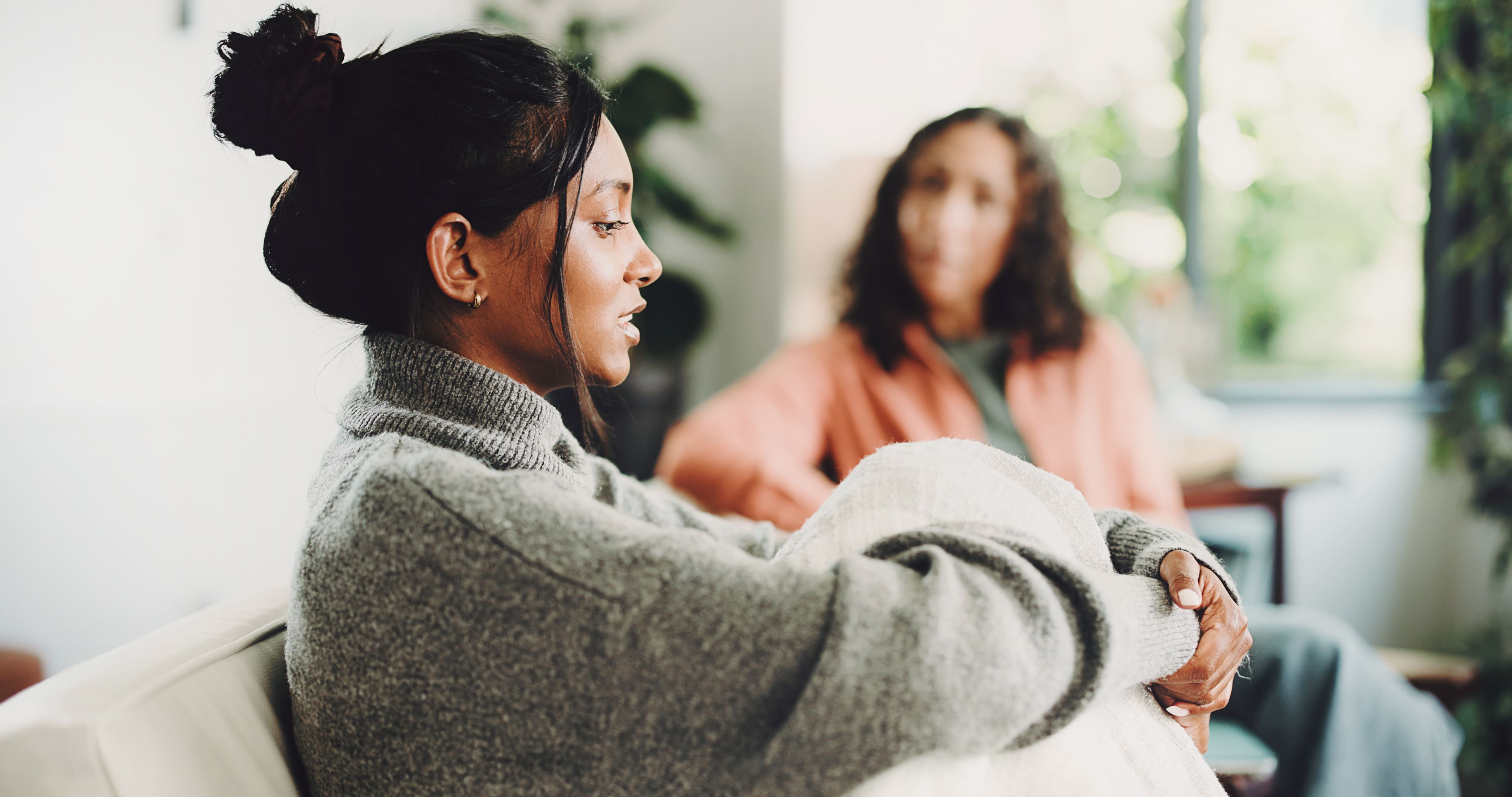 Woman in grey sweater sitting on sofa talking to another woman out of focus