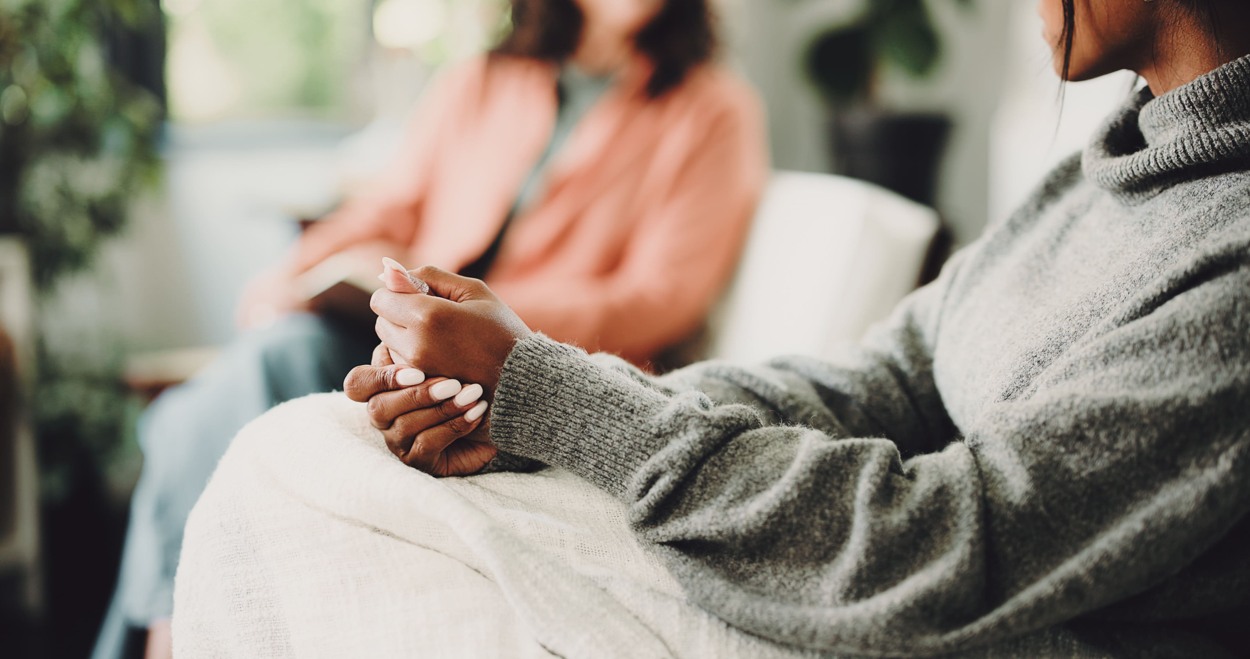 Woman wearing grey sweater sitting with hands clasped on lap, blurred person in background