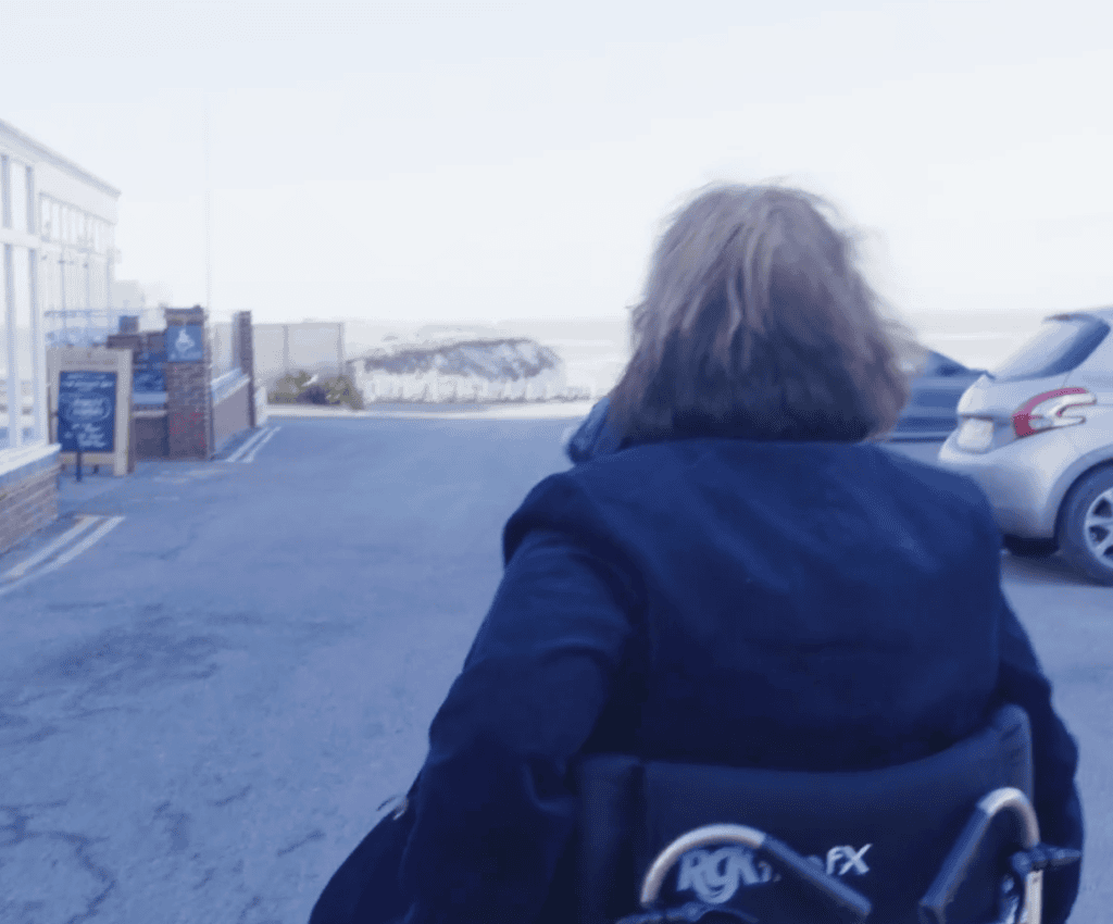 Person with shoulder-length hair in a wheelchair on a street near parked cars and buildings