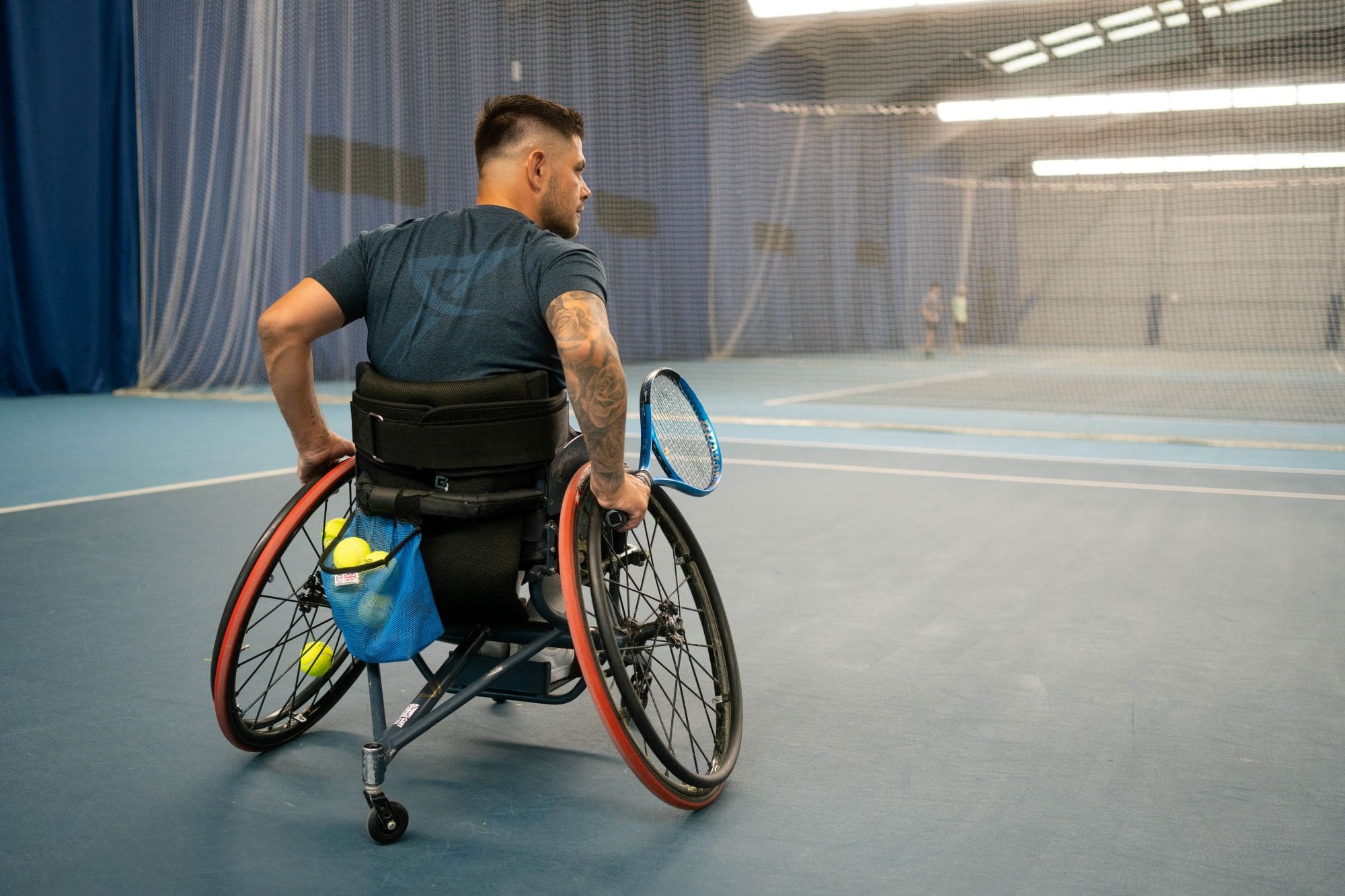 Man with tattooed arm in wheelchair holding a tennis racket on an indoor court
