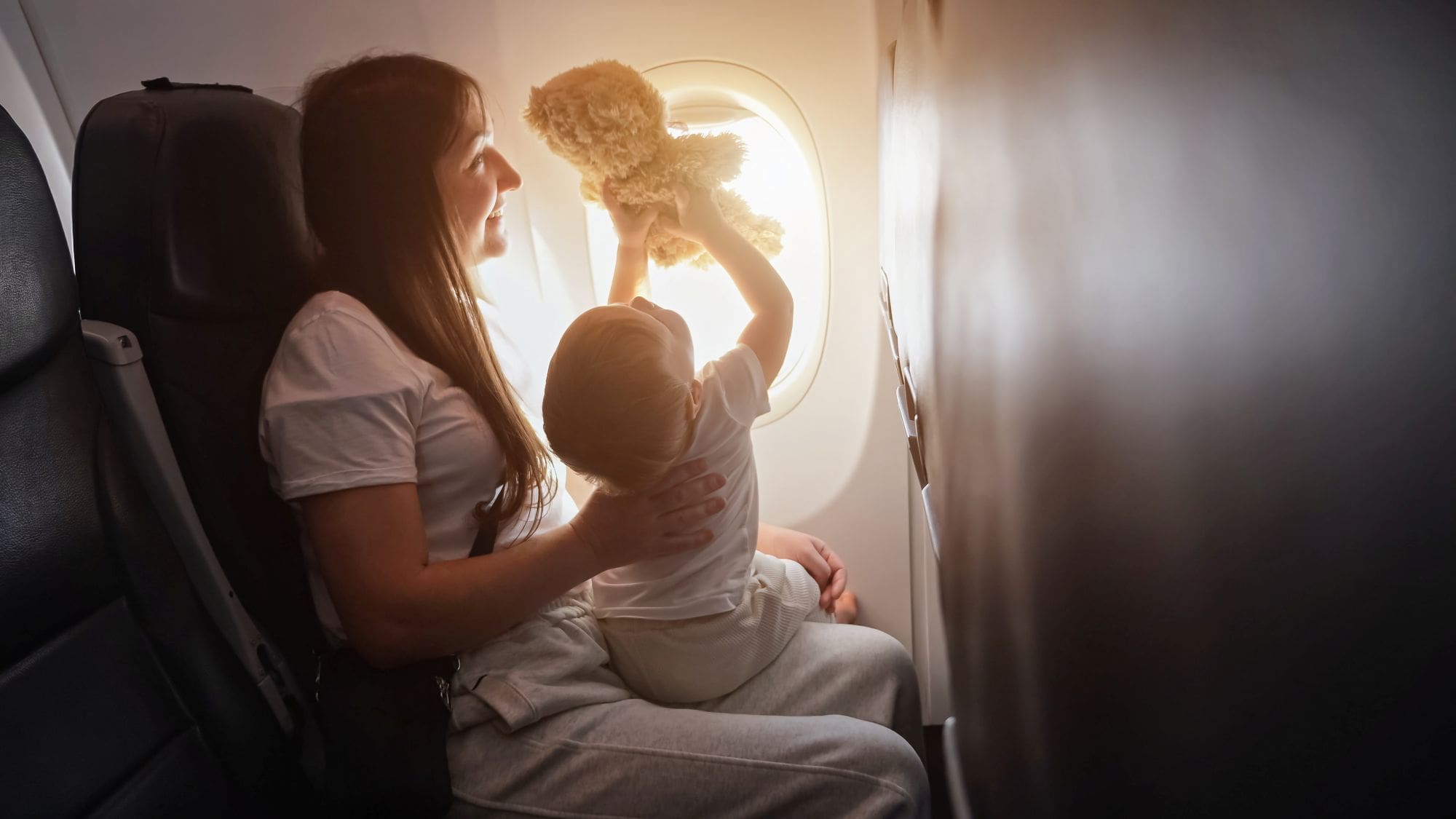 Woman and child playing with teddy bear near airplane window with sunlight