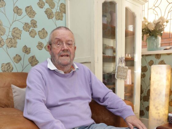 Older man wearing glasses and purple sweater sitting on brown leather sofa indoors
