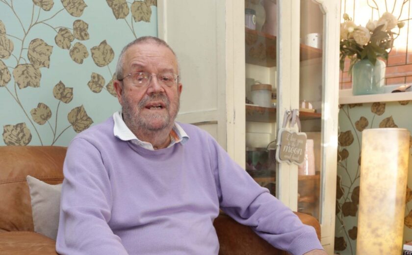 Older man wearing glasses and purple sweater sitting on brown leather sofa indoors