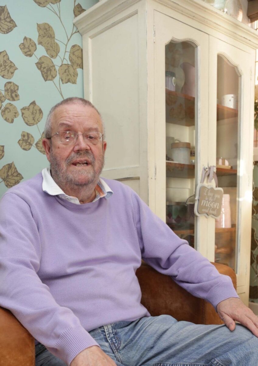 Older man wearing glasses and purple sweater sitting on brown leather sofa indoors