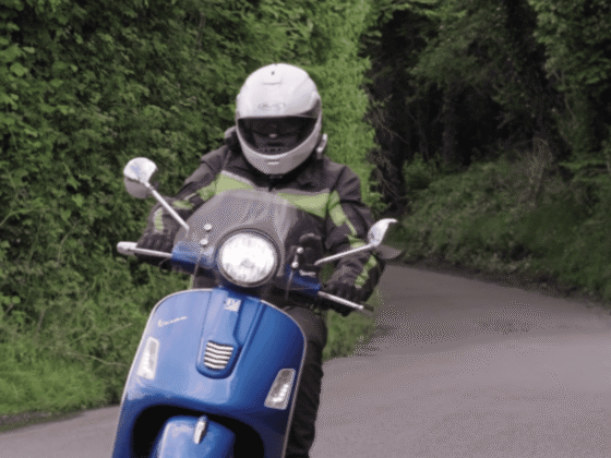 Person wearing helmet and protective clothing riding blue motor scooter on narrow road