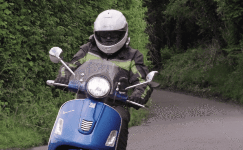 Person wearing helmet and protective clothing riding blue motor scooter on narrow road