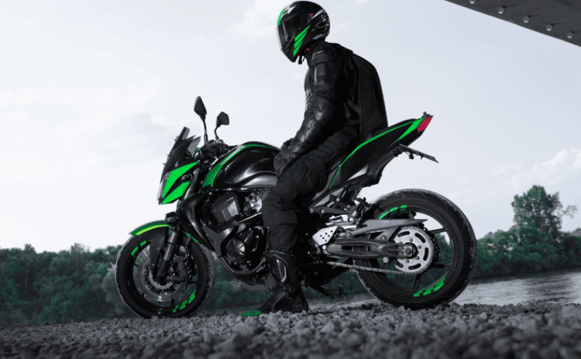 Motorcyclist in black gear and helmet sitting on a black and green motorcycle near a river under a structure