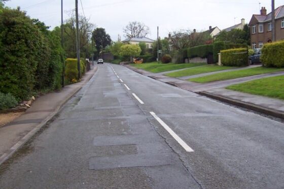 Empty residential street lined with houses and grassy gardens under cloudy sky
