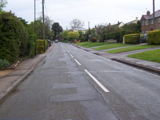 Empty residential street lined with houses and grassy gardens under cloudy sky