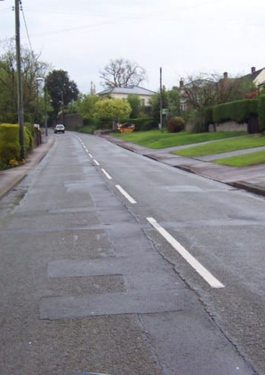 Empty residential street lined with houses and grassy gardens under cloudy sky