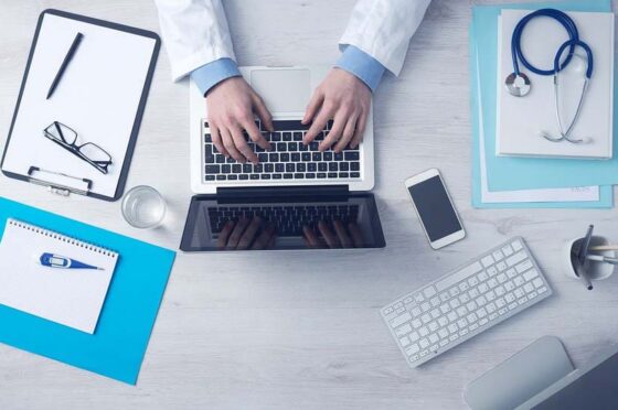 Overhead view of person in lab coat typing on laptop surrounded by medical documents and gadgets