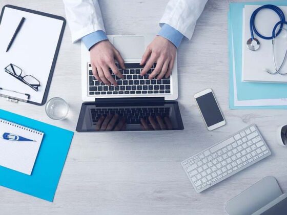 Overhead view of person in lab coat typing on laptop surrounded by medical documents and gadgets