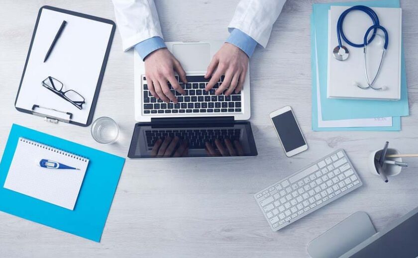 Overhead view of person in lab coat typing on laptop surrounded by medical documents and gadgets