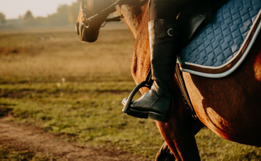 Close-up of horseback rider's boot in stirrup with horse and field at sunset
