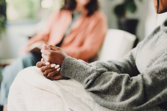 Woman wearing grey sweater sitting with hands clasped on lap, blurred person in background