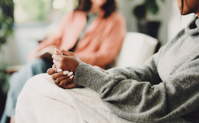 Woman wearing grey sweater sitting with hands clasped on lap, blurred person in background