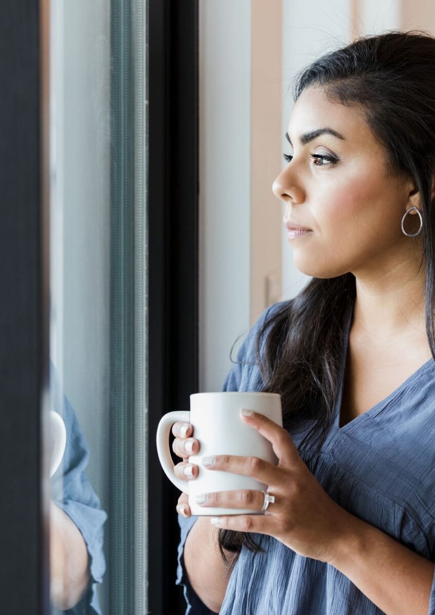 Mid adult woman stands at window with hot drink
